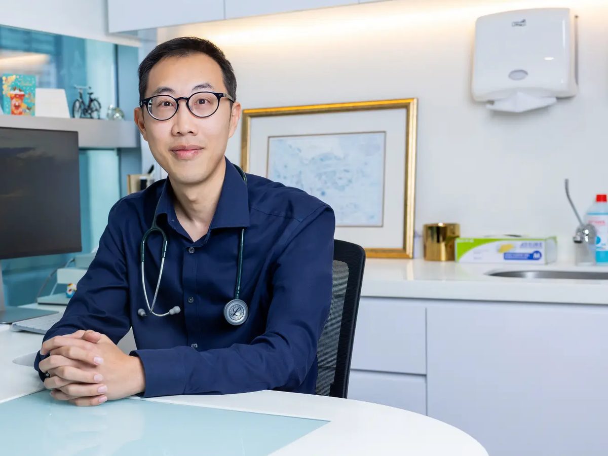 A man in round glasses and a navy shirt, with a stethoscope around his neck, at a desk in a consulting room